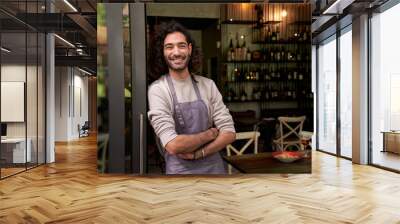 Portrait of happy young adult local small business owner looking at camera. Happy male bartender crossing his arms with positive attitude. Cheerful worker smiles while standing leaning on bar door. Wall mural