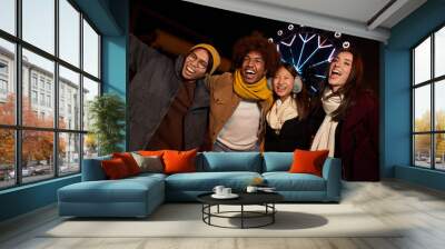 Portrait of mixed race group of happy colleagues having fun in winter clothes. Young smiling people laughing together enjoy the community. Happy face of young students at the amusement park at night. Wall mural