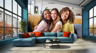 Portrait of three attractive female room mates looking smiling at camera. Group of cute cheerful girls hugging sitting on couch at home. Happy friends posing for photo indoors. Generation z and rent. Wall mural