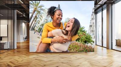 Two multiracial best friends having fun outdoor in summer. Caucasian woman walks piggyback African girl down street in city. Female enjoying their love relationship looking at each other and laughing Wall mural