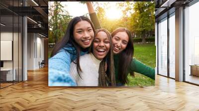 Vertical Portrait of three girls outside having fun taking selfie. An Asian Chinese woman, a black African American and a Caucasian together and embracing. Friendship in multi-ethnic groups of people Wall mural