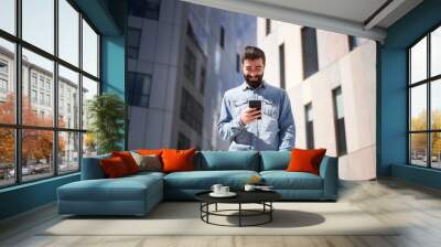 Young businessman in denim shirt using smart phone near office buildings in city financial center. Wall mural