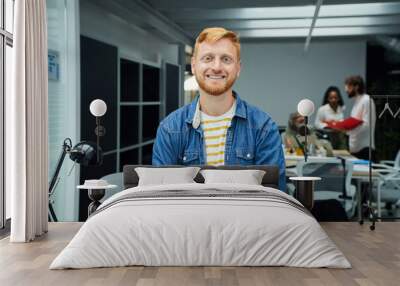 Young red-haired programmer or businessman in a modern office looking camera and smiles. Self-employed guy with his arms crossed poses for a photo in the meeting room. Colleagues work in background. Wall mural