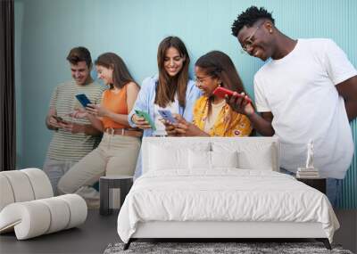 Young smiling multicultural group of friends using cell phones. Students leaning against a blue wall outside. Cheerful people gathered together looking at and typing on their mobiles.  Wall mural