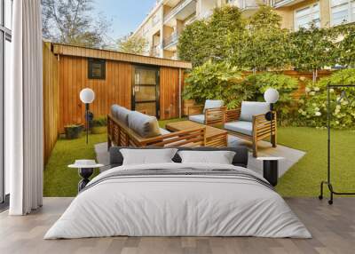 a backyard area with wooden furniture and green grass on the ground in front of an apartment building that is surrounded by trees Wall mural