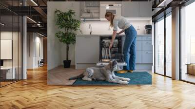 Woman loading dirty dishes in dishwasher and her dog lying on floor Wall mural