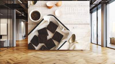 Brownies in a white tray with baking paper, egg shells, chocolate in a white bowl, two spoons and a white napkin, on a white wooden backdrop.  Wall mural