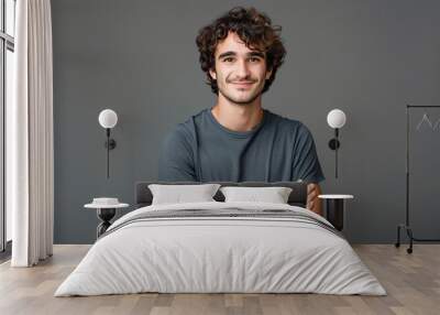 Photo of a happy young man with curly hair in a gray shirt standing on a gray background with his arms crossed and looking at the camera smiling Wall mural