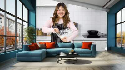 A happy woman in an apron, mixing batter in a clear glass bowl with a wooden spatula. Standing in a clean and modern kitchen Wall mural