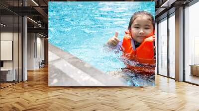 Asian child playing in the pool. Wearing orange life jacket, smiling with thumbs up. Wall mural