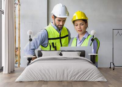 Young engineer with laptop computer explain to foreman about a floor plan. Both wear construction helmet and safety vest. Work environment at the construction site of housing projects. Wall mural