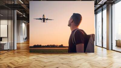 Man with backpack looking up to airplane landing at airport during beautiful summer sunset. . Wall mural