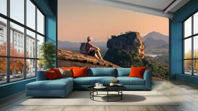 Man with backpack sitting on rock and looking at landscape at sunset. Beautiful scenery with Sigiriya rock. Solo traveler in Sri Lanka. . Wall mural