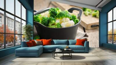 Black saucepan with cooked broccoli and cauliflower on wooden kitchen table. Wall mural