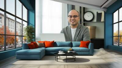 A middle-aged man around the age of 35. Working at home Work through the laptop. He was wearing a grey suit and glasses. Smiling asian businessman work from home.. Wall mural