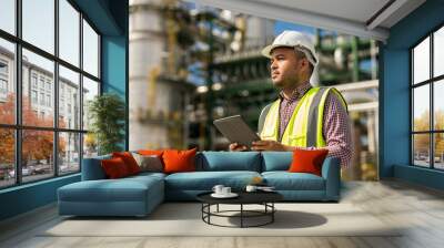 Asian engineer handsome man use tablet with white safety helmet standing front of oil refinery. Industry zone gas petrochemical. Factory oil storage tank and pipeline. Workers in a refinery Wall mural