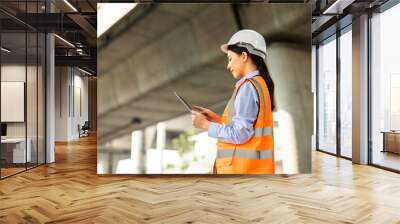 Asian engineer worker woman or architect looking construction with tablet wear white safety helmet in construction site. Standing at highway concrete road site. Progress planning of highway bridge. Wall mural