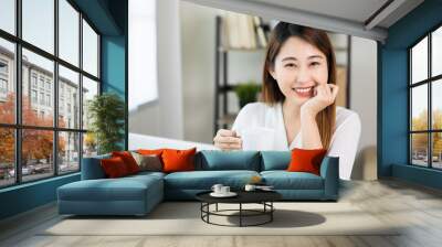 Young asian beautiful business woman working with laptop sitting at home drinking coffee. Smiling charming happy young female doing homework meeting conference with team at home. Wall mural