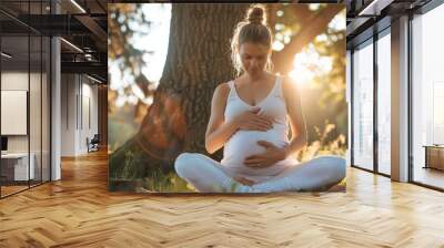 Portrait of white beautiful pregnant woman doing yoga exercise in park near tree, touching belly and heart Wall mural