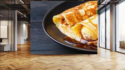 Delicious and golden crepes served on a dark plate, garnished with mint leaves and drizzled with syrup. Wall mural