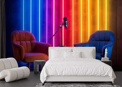 A vibrant podcast studio backdrop featuring two stylish chairs illuminated by neon lights in blue, pink, and yellow. atmosphere is energetic and inviting, perfect for engaging conversations Wall mural