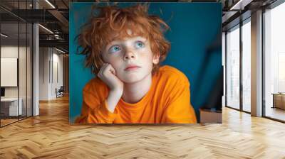 A thoughtful young boy with curly red hair gazes upward while resting his chin on his hand Wall mural