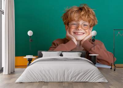 Cheerful boy with glasses studying at a wooden desk in a classroom, enjoying reading at midday Wall mural