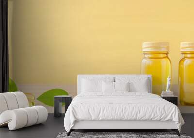 Two glass jars filled with golden oil alongside capsules and fresh leaves on a yellow background Wall mural