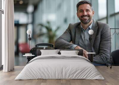 A cheerful man is seated with his sleek luggage in an airport lounge, clad in a formal blazer and shirt, embodying confidence and readiness for an impending journey. Wall mural