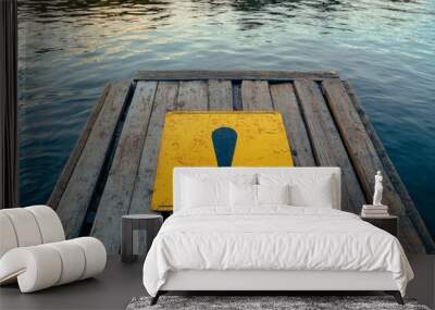 A yellow warning sign with an exclamation point is placed on a floating wooden dock over water, emphasizing awareness and safety amidst a serene, aquatic setting. Wall mural