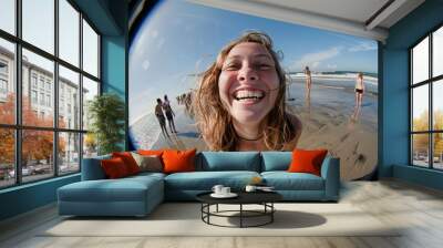 A young woman smiling happily at the camera on a sunny beach day with clear blue skies and other beachgoers and the ocean in the background. Wall mural