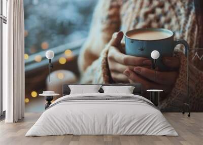 Hands holding a warm cup of coffee, surrounded by a cozy winter setting with warm lights and comfortable sweater, creating a sense of comfort and relaxation in a home environment. Wall mural