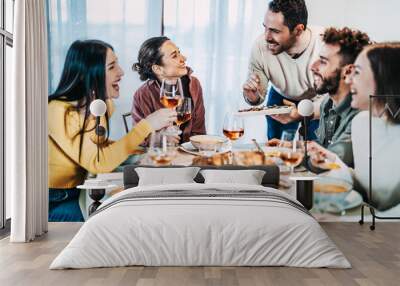 Happy group of friends having dinner party at home - Cheerful young people having lunch break together - Life style concept with guys and girls celebrating thanksgiving - Man serves vegetables Wall mural