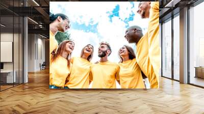 Hugging each other and smiling a group of volunteer people helping others - A coluoured team of Men and Women  from different ethnicities embrace in a circle - Joyful community lifestyle concept. Wall mural