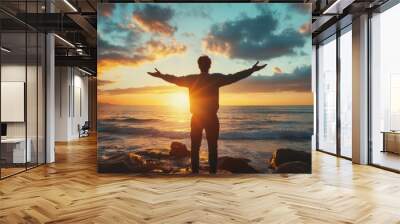 A man stands on a beach with his arms outstretched, looking up at the sky Wall mural