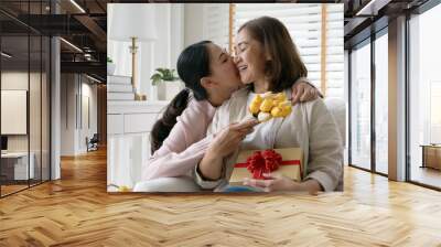 Attractive beautiful asian middle age mum sit with grown up daughter give gift box and flower in family moment celebrate mother day. Overjoy bonding cheerful kid embrace relationship with retired mom. Wall mural