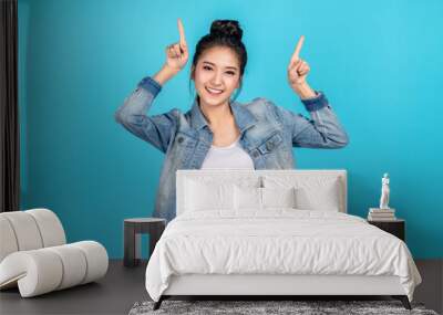 Happy asian woman feeling happiness and standing pointing hands up on blue background.Cute asia girl smiling wearing casual jeans shirt and finger pointing to above for present promotions Wall mural