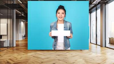 Happy asian woman standing and holding plus or add sign on blue background. Cute asia girl smiling wearing casual jeans shirt and showing join sign for increse, upgrade and more benefit concept Wall mural