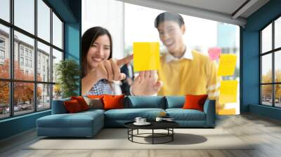 Young successful creative man and woman smile and brainstorm on project at office pointing hand or finger to yellow post note or sticky note. Close up view couple sharing idea on glass wall concept. Wall mural