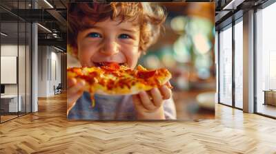 A little white boy with curly hair with a smile on his face is eating a piece of delicious hot pizza. The pizza is covered with pepperoni and cheese. Pizza day. Italian cuisine Wall mural