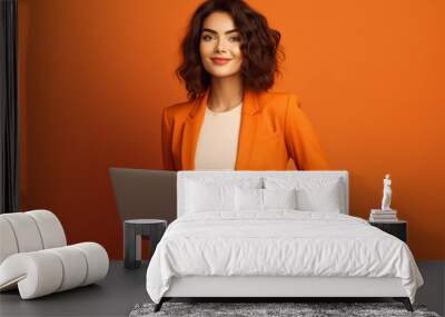 Smiling business woman wearing orange business suit with a laptop at armchair, isolated on orange background Wall mural