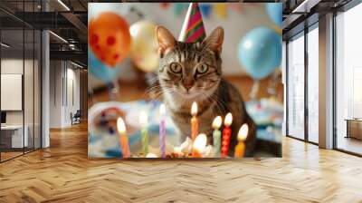 A cat wearing a party hat sits behind a birthday cake with lit candles, surrounded by colorful balloons on a festive background, celebrating a birthday. Wall mural