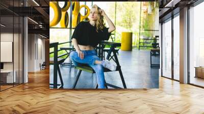 A young woman sits on a chair in a cafe in a relaxed pose. A girl in sportswear. Wall mural