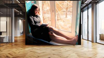 perfect young brunette girl in a long black dress sits on a windowsill by the window and reads a book Wall mural