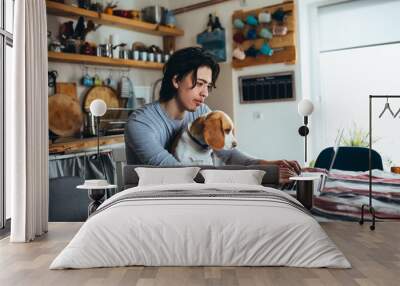 young man holding his dog in nap while using laptop in kitchen Wall mural