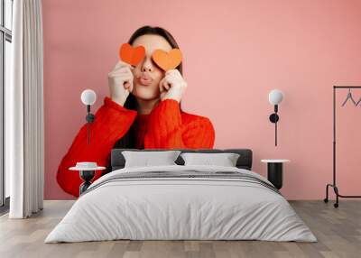 Cute girl is sending a kiss and covering her face with paper hearts. The girl is standing isolated against the pink background. Concept of the St. Valentine's Day Wall mural