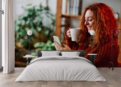 A cute ginger girl with curly hair is sitting in the chair at home in the morning, texting messages on the phone and drinking her coffee. Wall mural