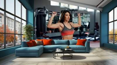 A fit, strong female bodybuilder is lifting dumbbells and doing exercises for biceps in a gym. Wall mural