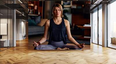 A middle-aged woman sitting in a lotus yoga pose and meditating with eyes closed. Wall mural