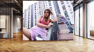 An urban teenage girl is sitting on the railing in the urban exterior while her boyfriend is in the background looking around. Wall mural